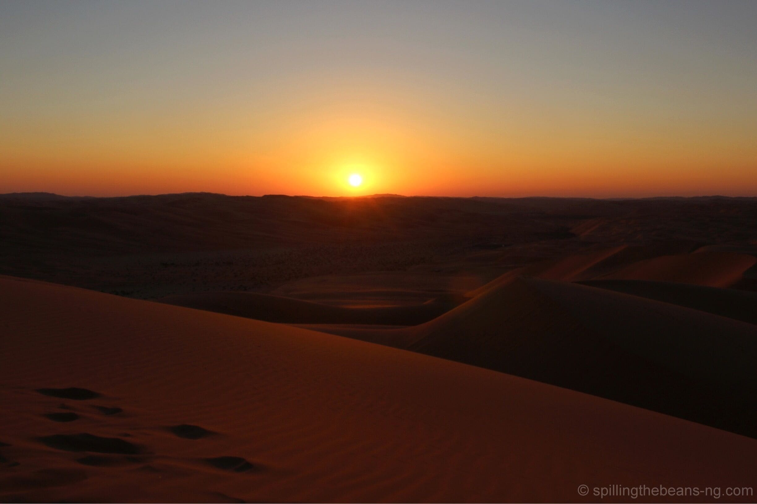 A desert landscape at sunset with sand dunes and a visible sun.