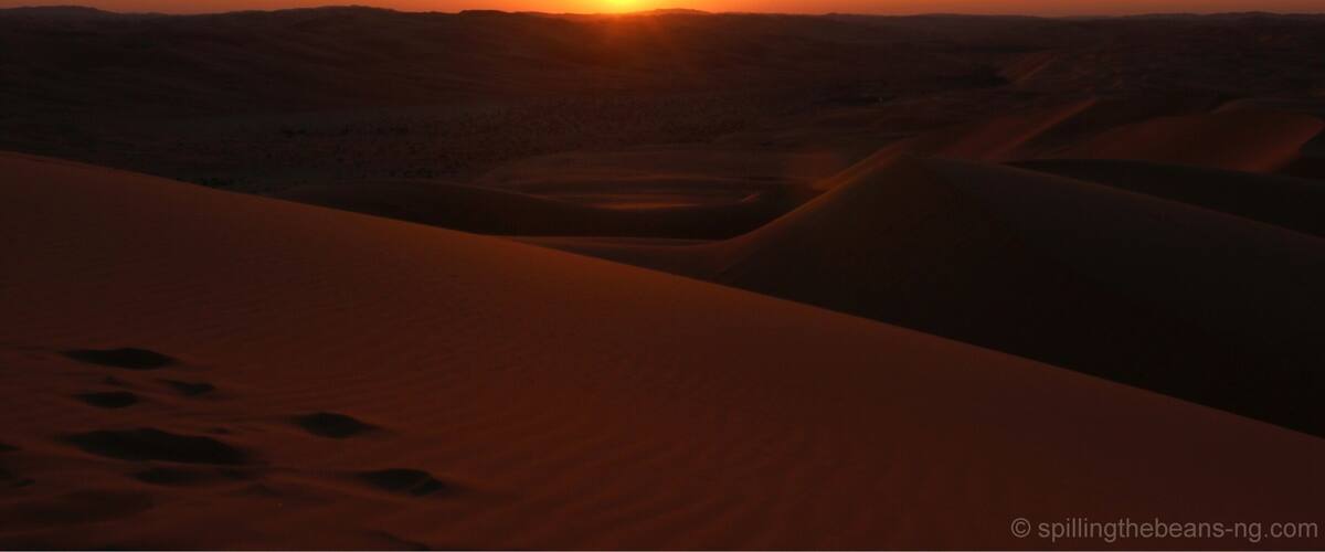 A desert landscape at sunset with sand dunes and a visible sun.