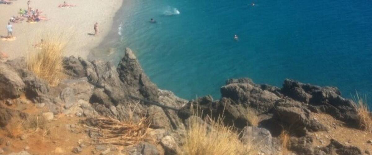 A rocky coastline with a beach, clear blue sea, and distant mountains.
