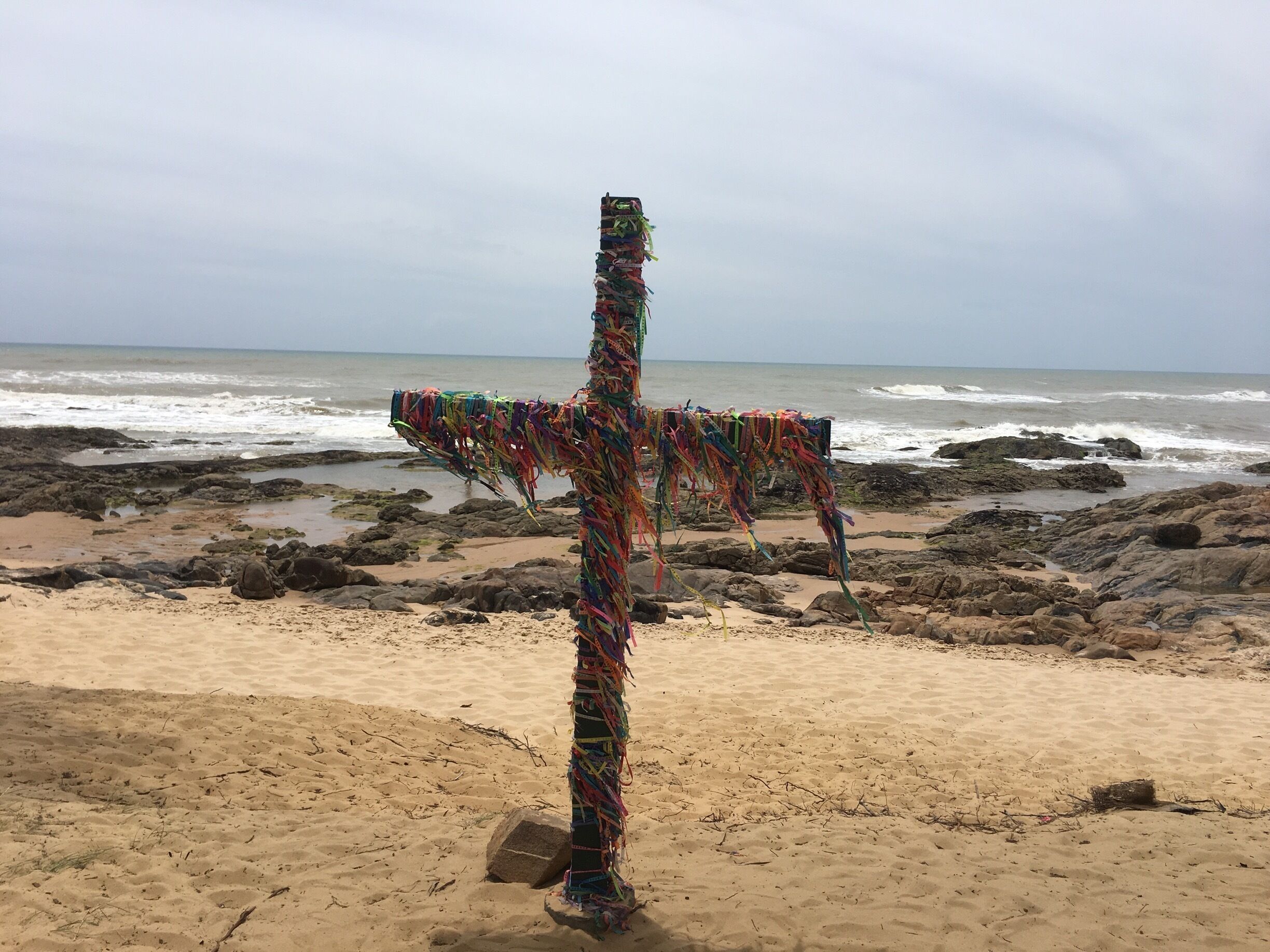 A colorful cross on a rocky beach with waves in the background.