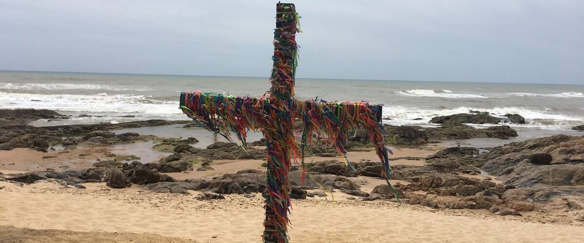 A colorful cross on a rocky beach with waves in the background.