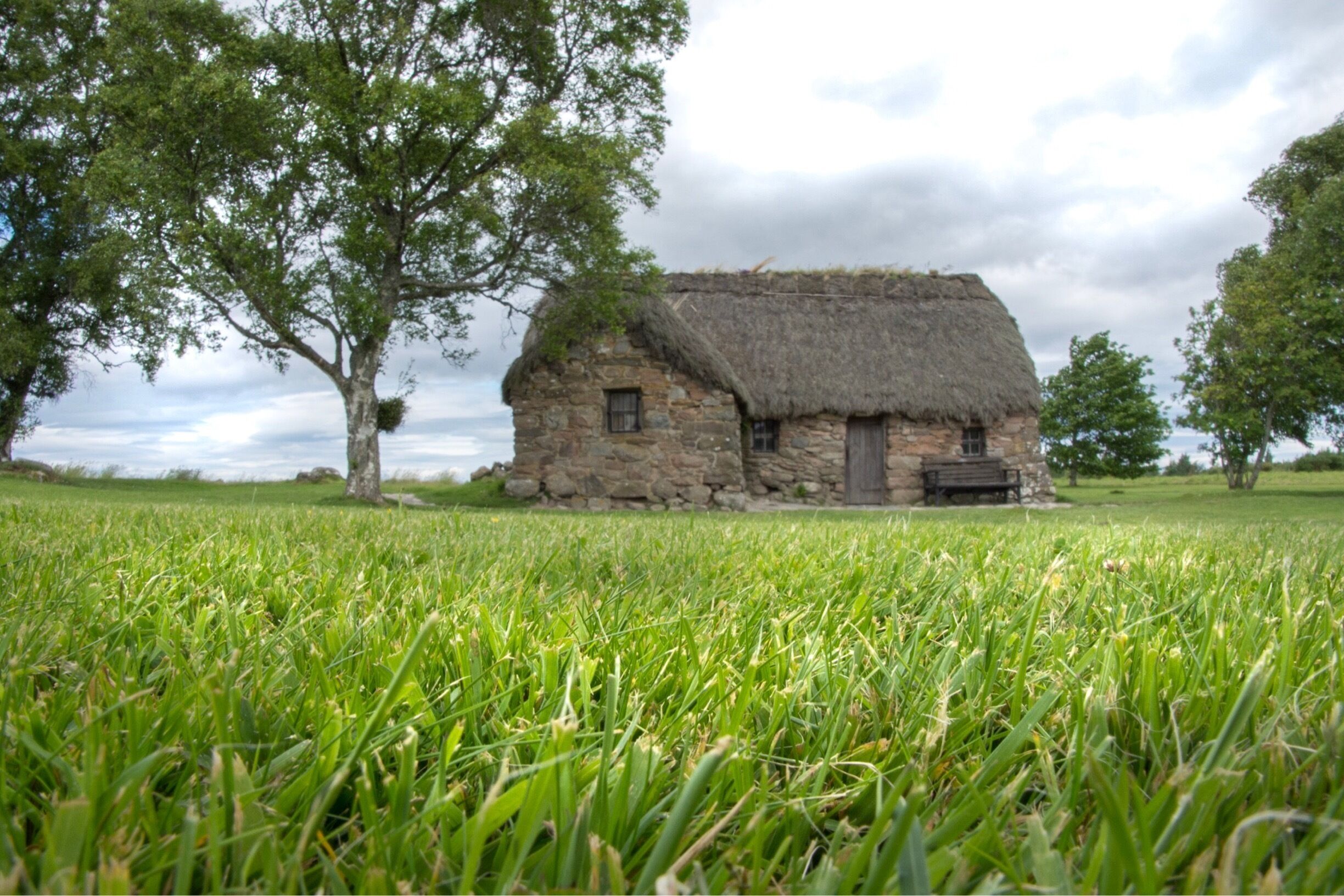 A stone house with a thatched roof surrounded by lush green grass and trees.