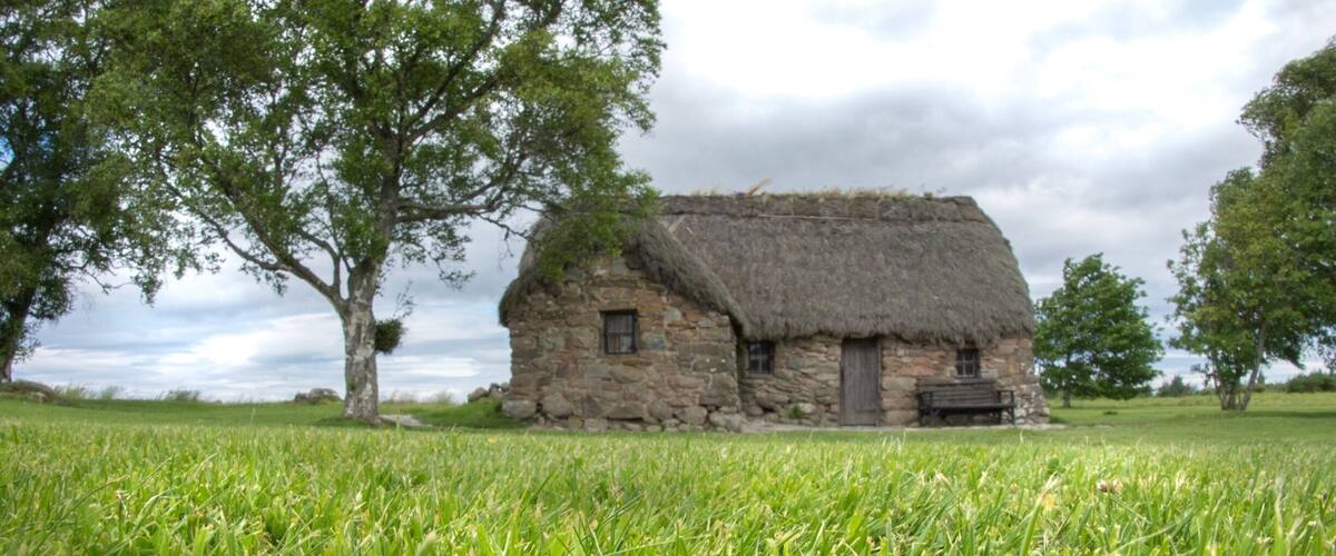 A stone house with a thatched roof surrounded by lush green grass and trees.