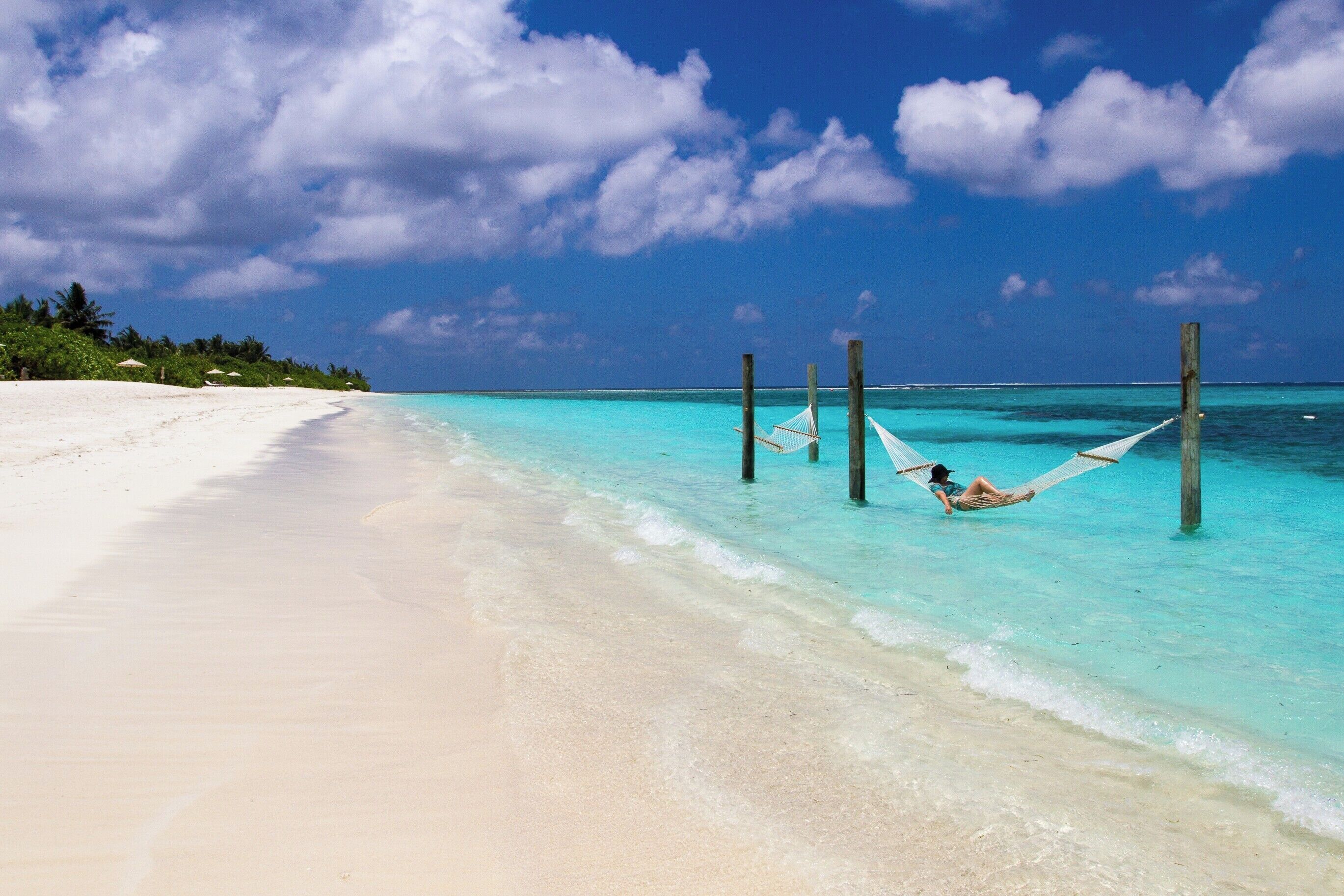 A beach with clear turquoise water, white sand, and a hammock with a person lying in it.