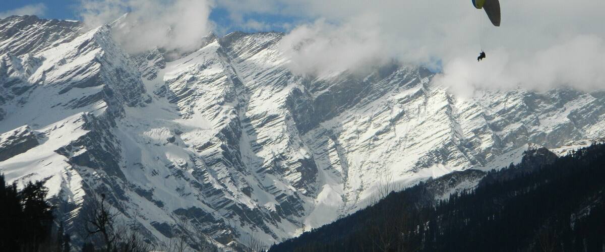 A paraglider flying over snow-covered mountains with a forested valley below.