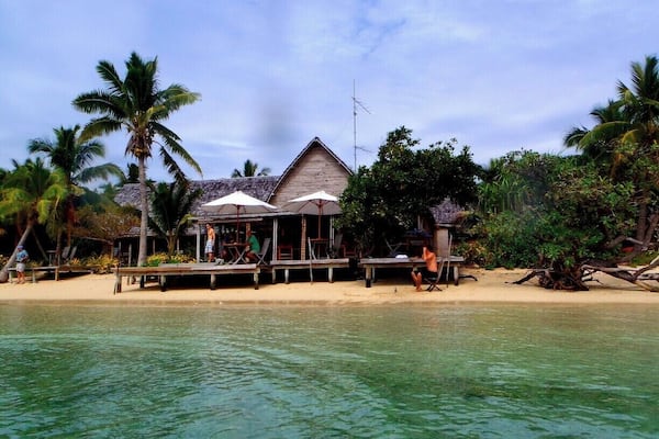 A beachside resort with a thatched-roof building, palm trees, and clear turquoise water.
