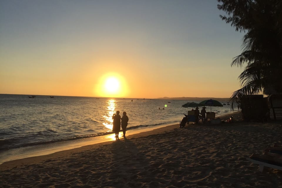 A beach at sunset with the sun low on the horizon, people walking along the shore, and a few boats on the water.