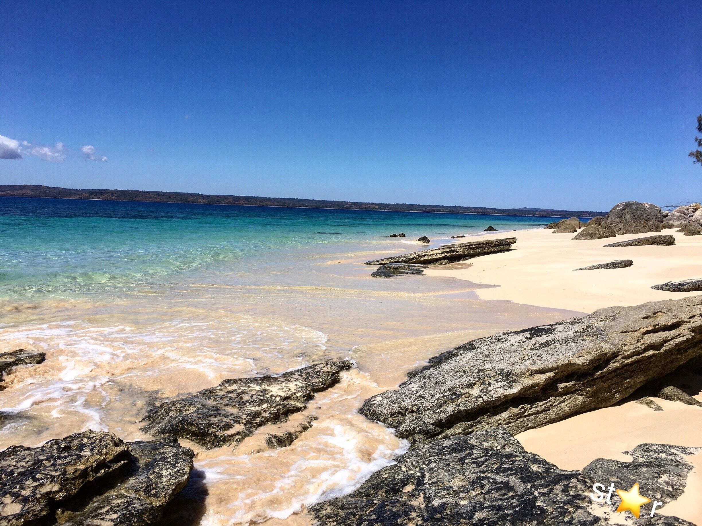 A beach with clear turquoise water, rocky shoreline, and a distant hilly landscape.