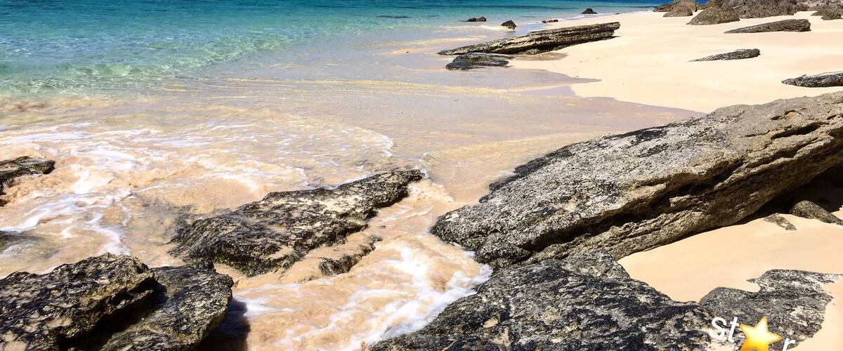 A beach with clear turquoise water, rocky shoreline, and a distant hilly landscape.