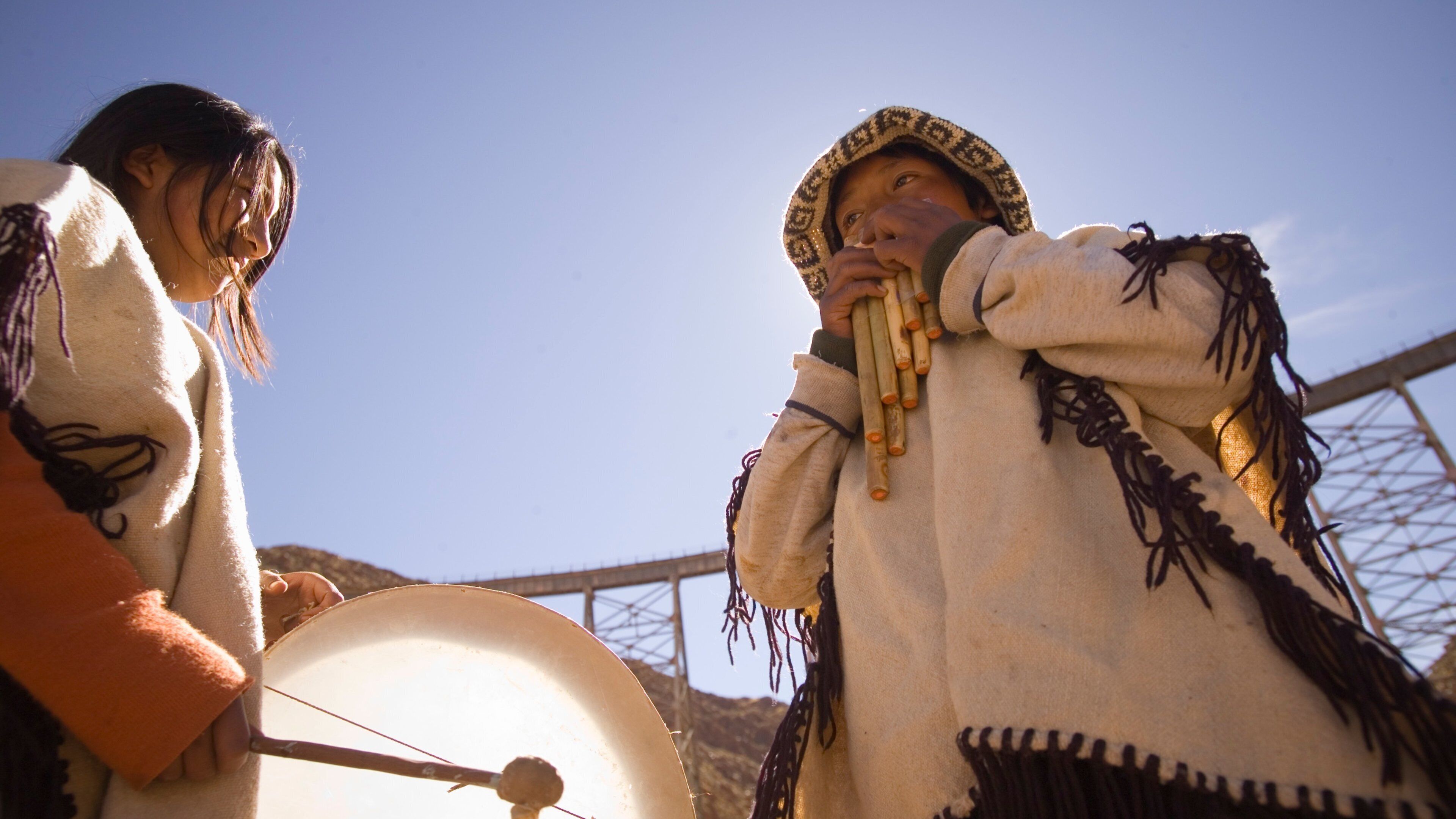 Two individuals in traditional attire, one playing a drum and the other holding a flute, outdoors with a clear sky and a bridge structure in the background.