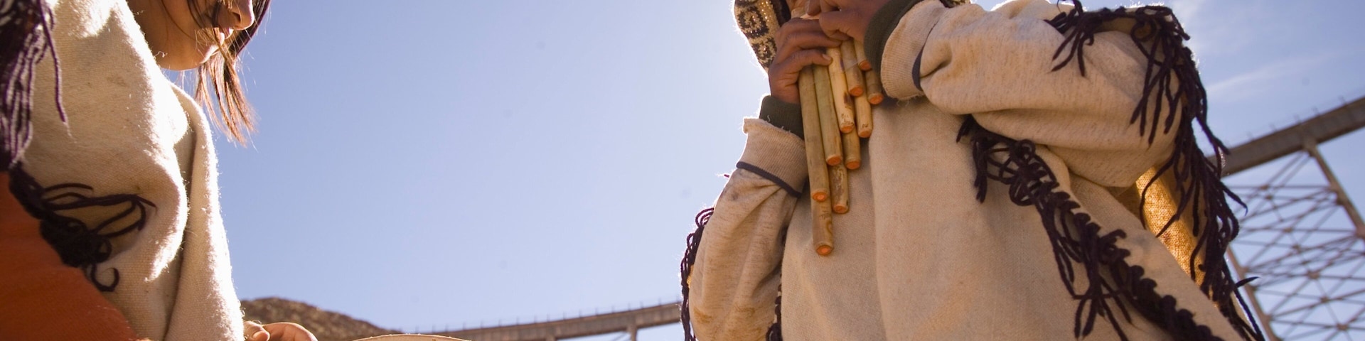Two individuals in traditional attire, one playing a drum and the other holding a flute, outdoors with a clear sky and a bridge structure in the background.