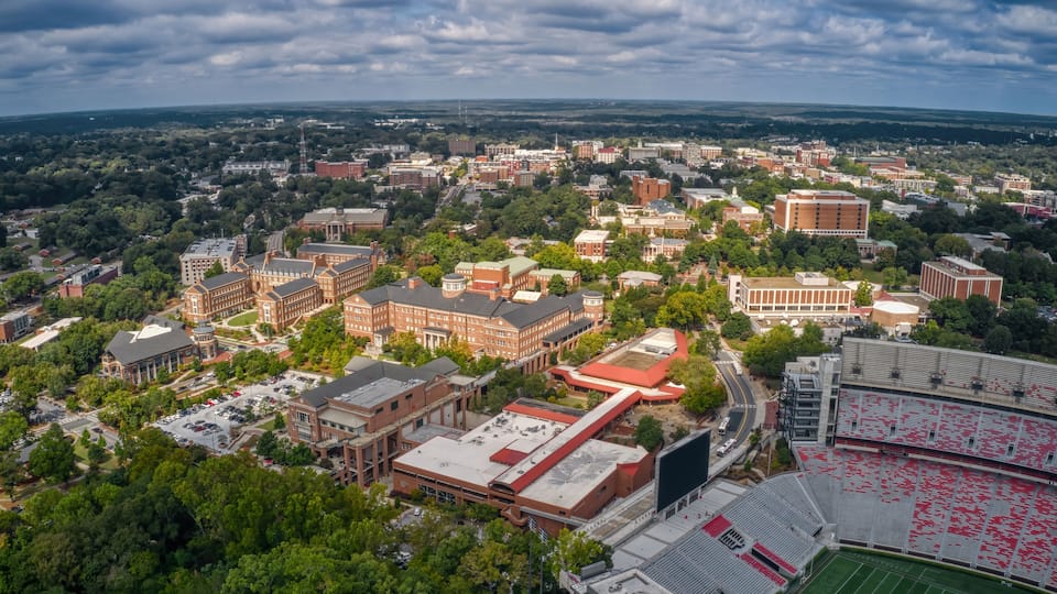 An aerial view of a university campus with multiple buildings, a stadium, and surrounding greenery.