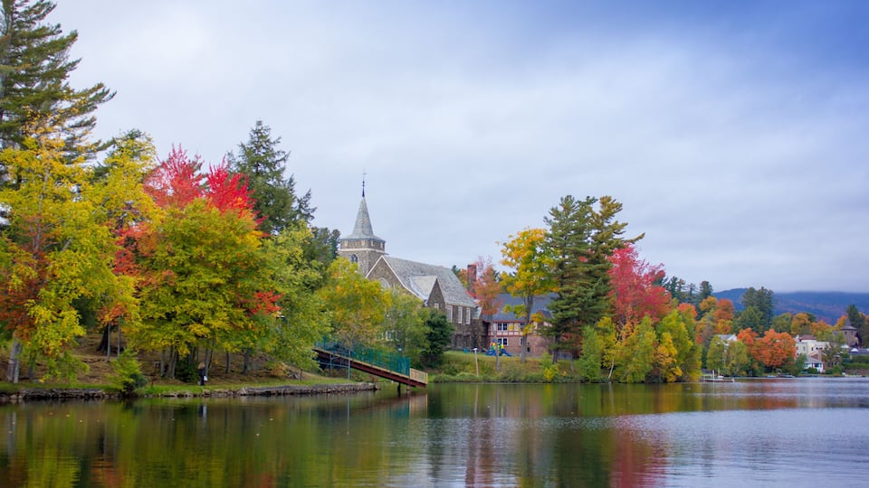 A serene lake with autumn foliage, a church with a steeple, and a small bridge.