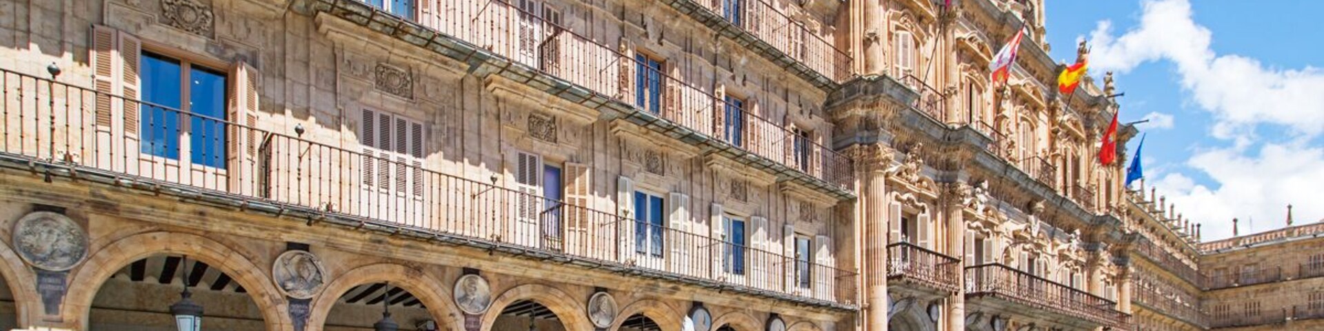 Un edificio histórico con arquitectura ornamentada, un patio con mesas al aire libre y banderas.