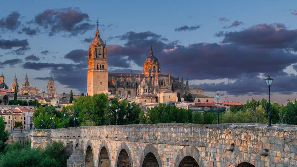 A historic stone bridge with arches, a prominent church with a tall bell tower, and a cityscape with various buildings under a twilight sky.