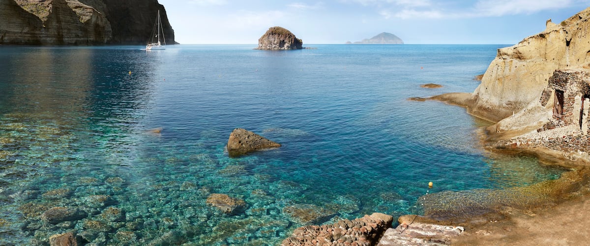 A coastal landscape with clear blue waters, rocky shores, and a sailboat.