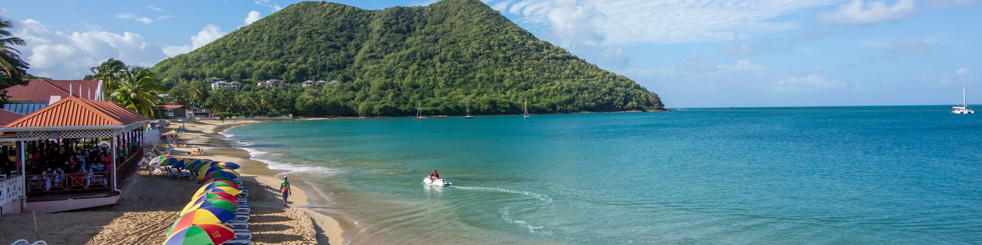 Uma praia com uma fileira de guarda-sóis coloridos, mar azul límpido e uma colina verde exuberante ao fundo.