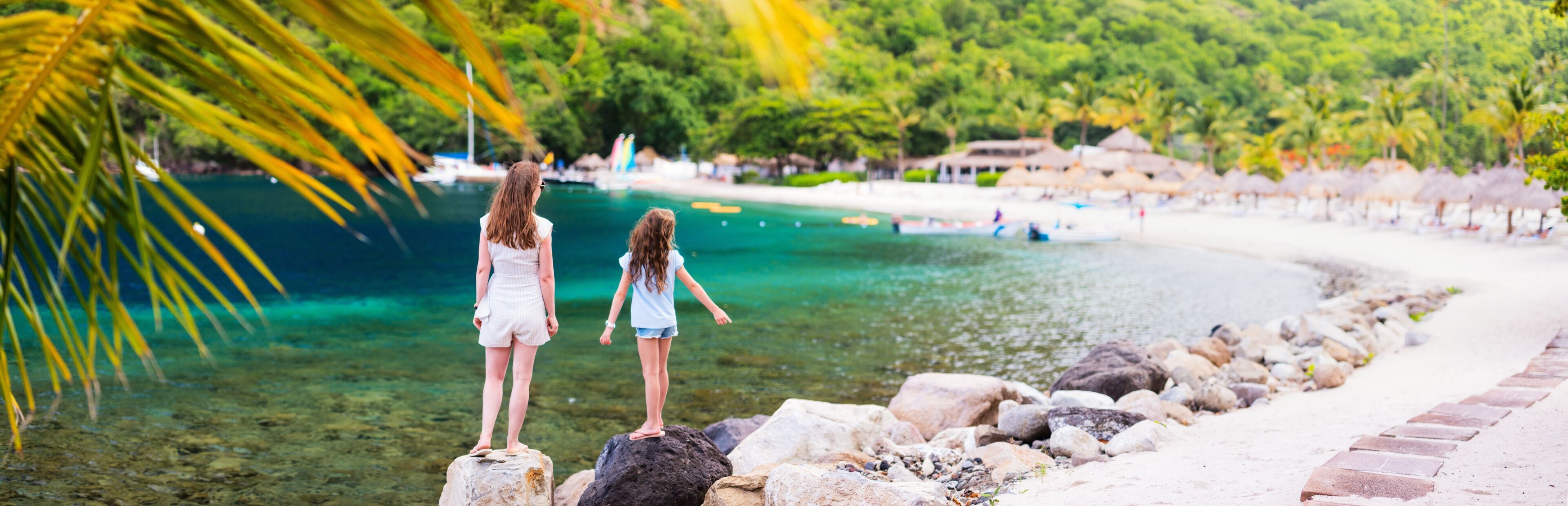 Dos personas de pie sobre rocas junto a un mar azul y cristalino, con una playa y palmeras al fondo.
