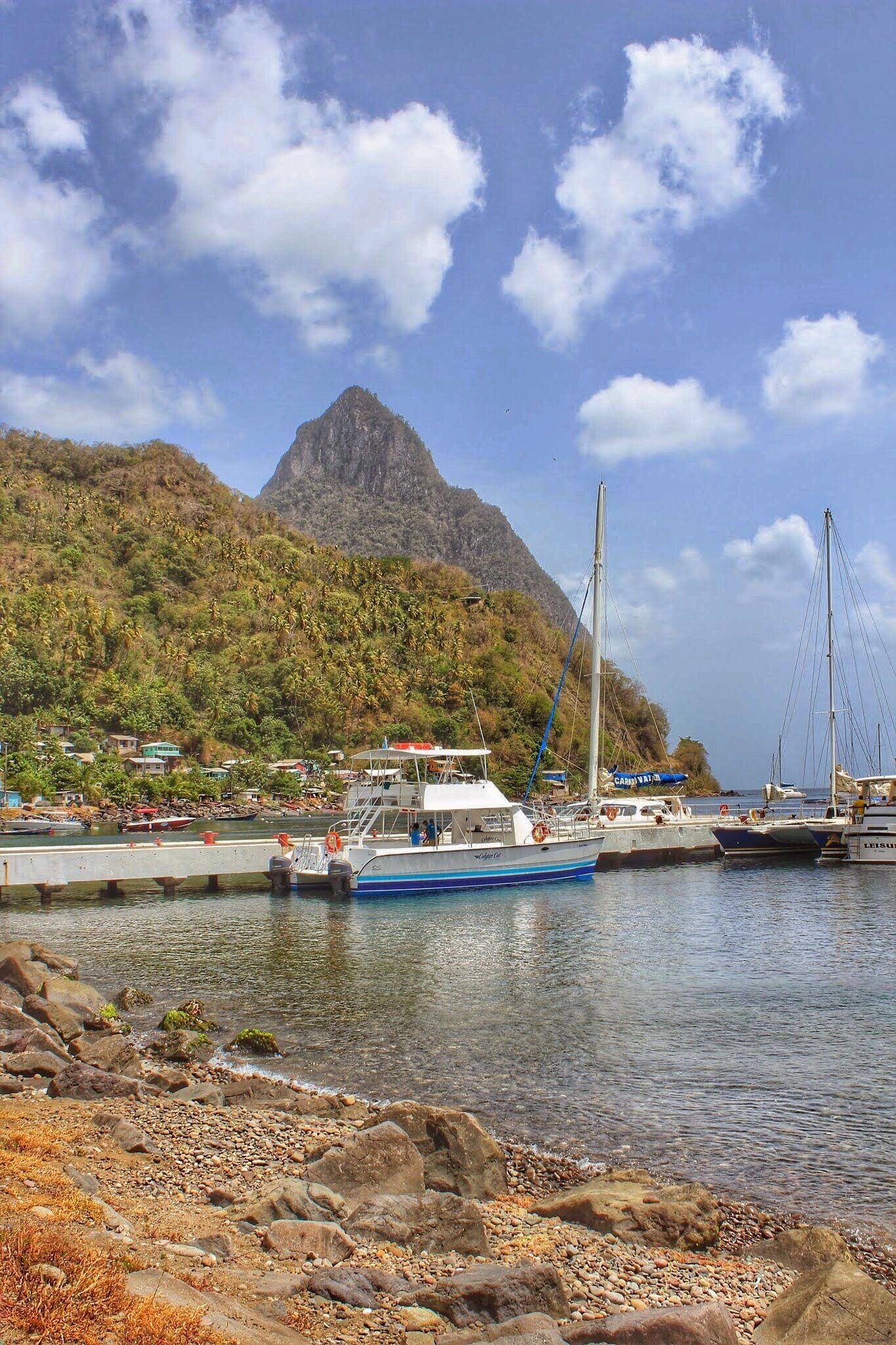 A coastal scene with boats docked at a pier, a mountain in the background, and a rocky shoreline.