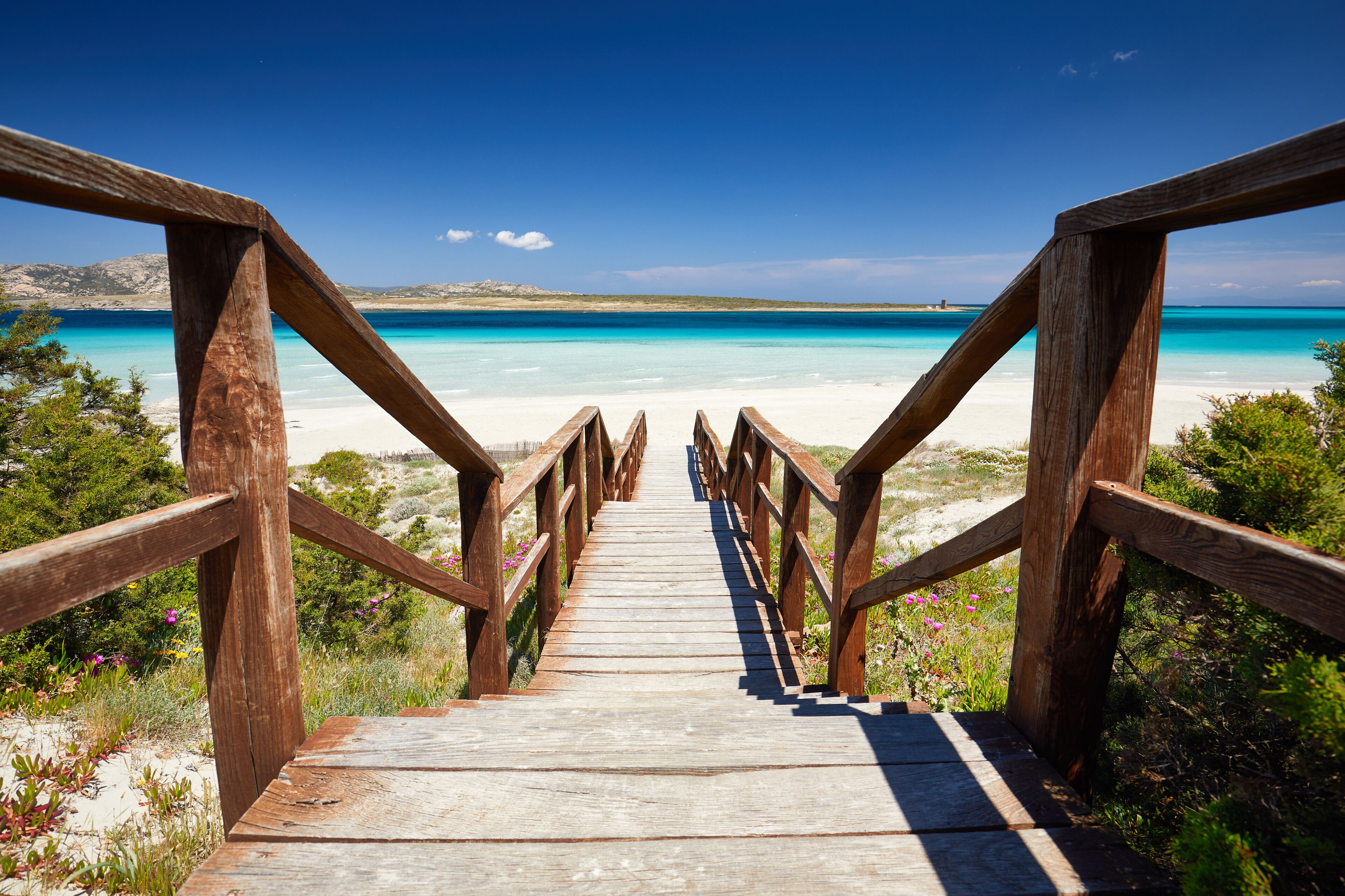 Una escalera de madera que conduce a una playa con aguas turquesas cristalinas y montañas lejanas.