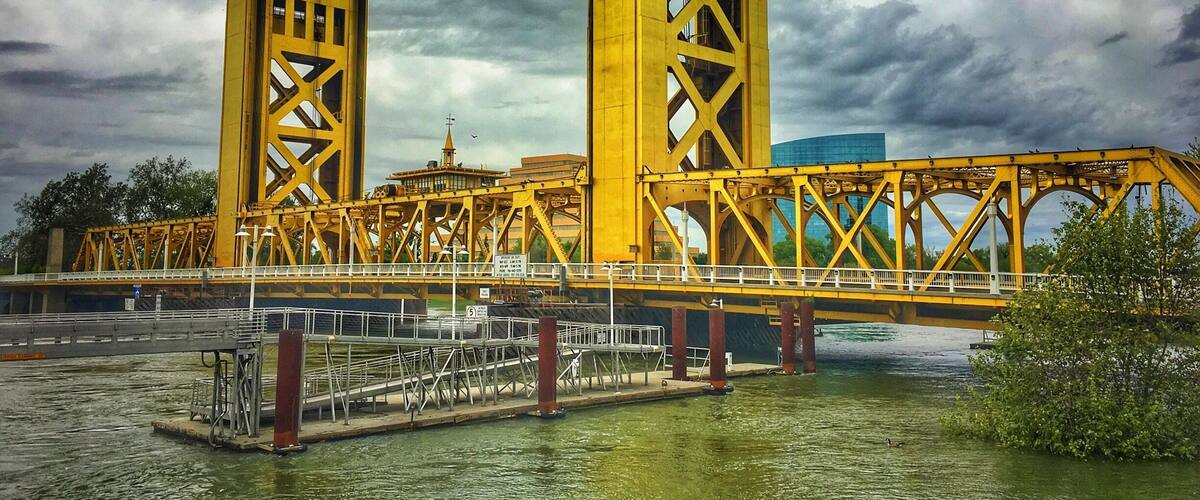 A yellow bridge with a complex truss design over a body of water, with a cloudy sky in the background.
