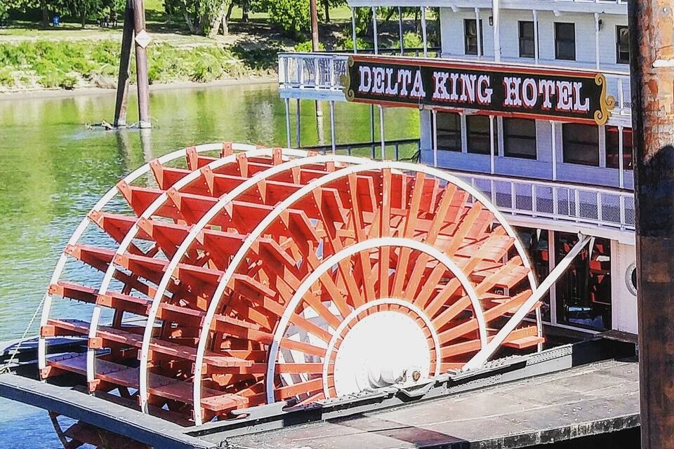 Um barco a vapor com uma grande roda de pás vermelha atracado próximo a um hotel chamado "Delta King Hotel" às margens de um rio.