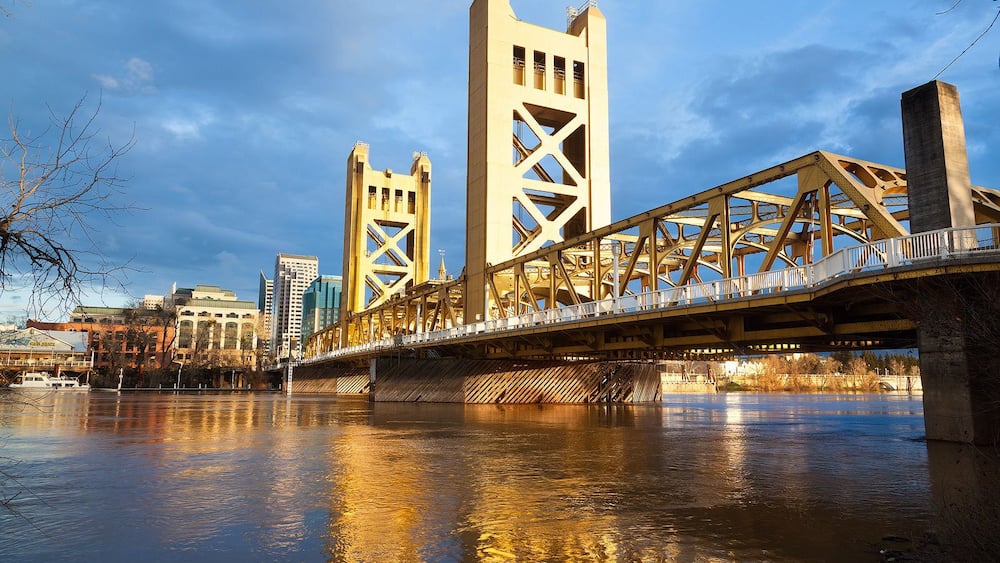 A bridge with a unique lattice structure spanning a river, with buildings in the background.