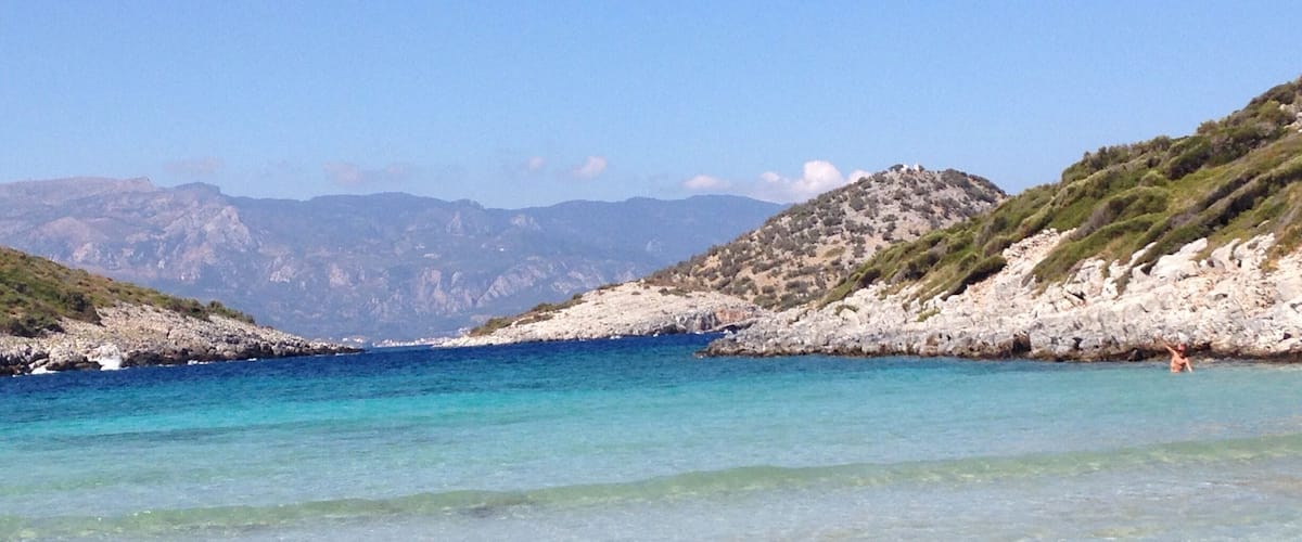 A coastal landscape with clear turquoise waters, rocky hills, and a distant mountain range.