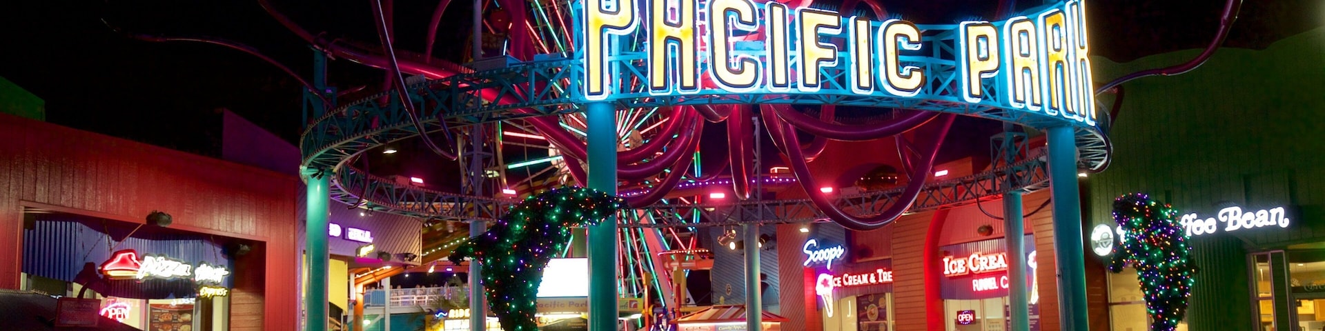 Un parque de atracciones muy iluminado con un letrero de neón que dice "PACIFIC PARK" y diversas tiendas y atracciones.