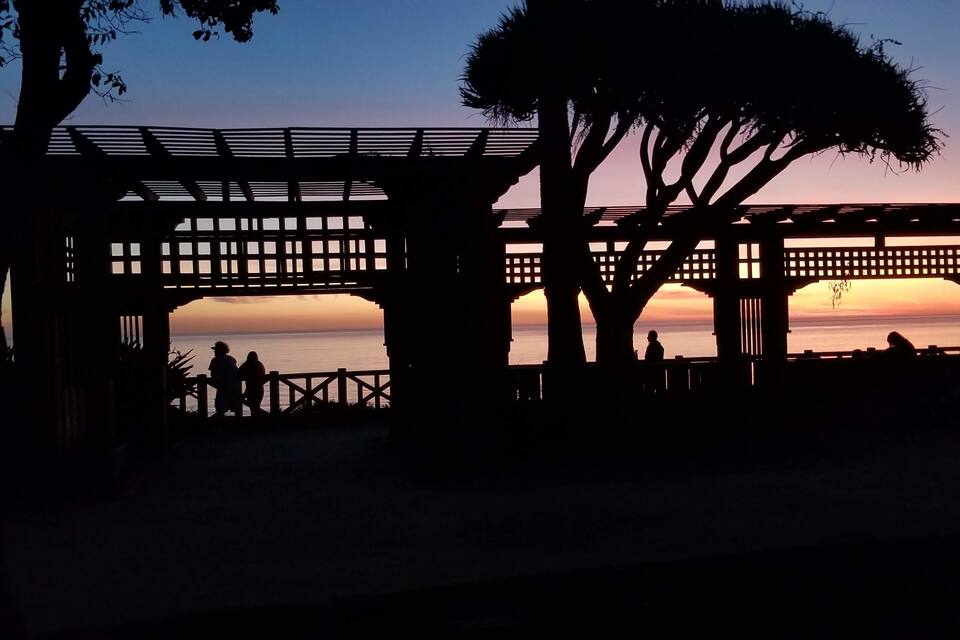 A sunset view through a wooden pergola with people silhouetted against the sky.