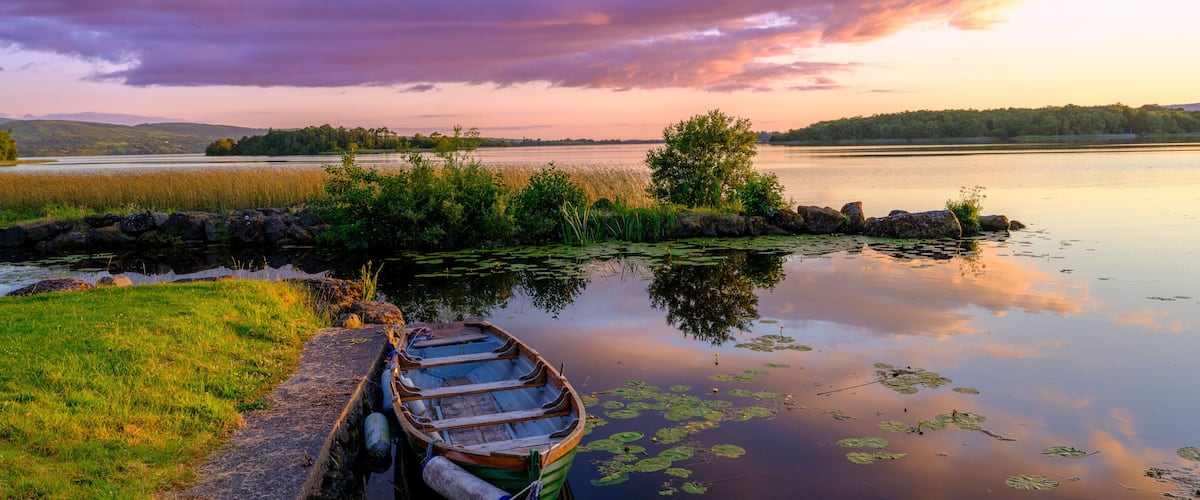 A calm lake with a wooden boat docked on the left, surrounded by greenery and a stone wall.