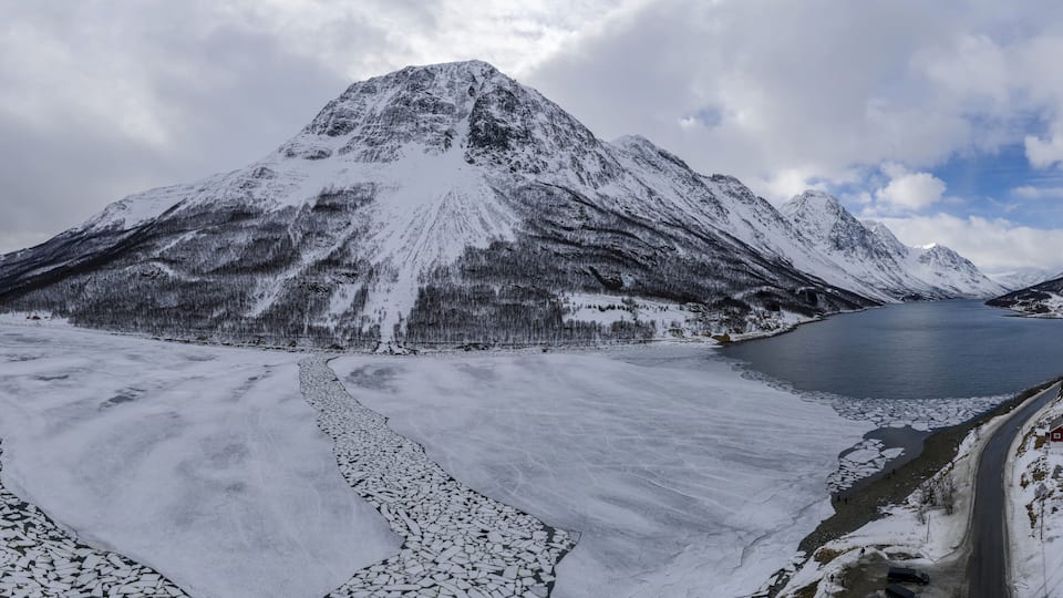 A snow-covered mountain range with a frozen lake and a road leading to a small settlement.