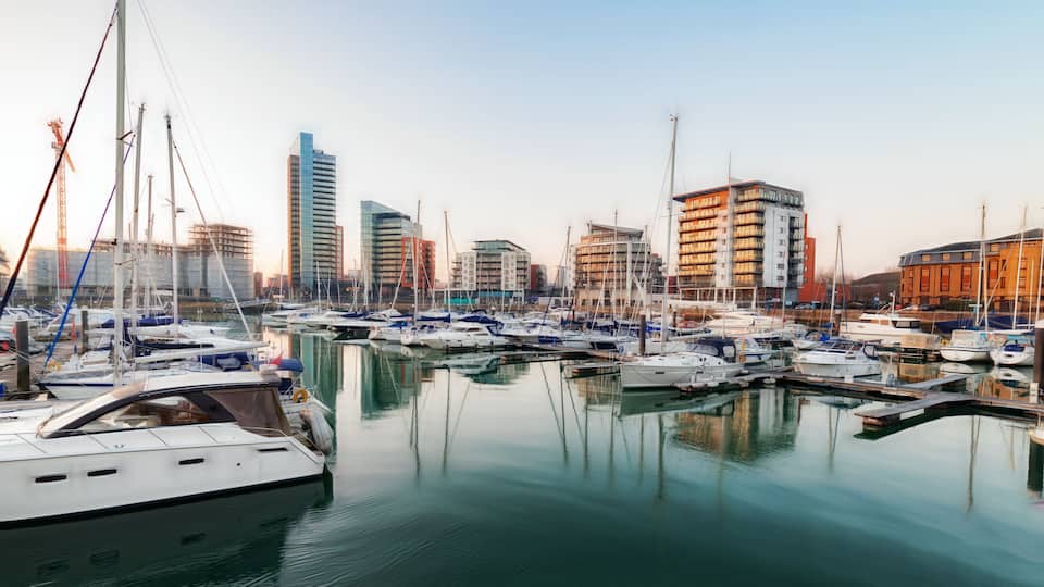 A marina with multiple boats docked, surrounded by modern buildings and cranes.
