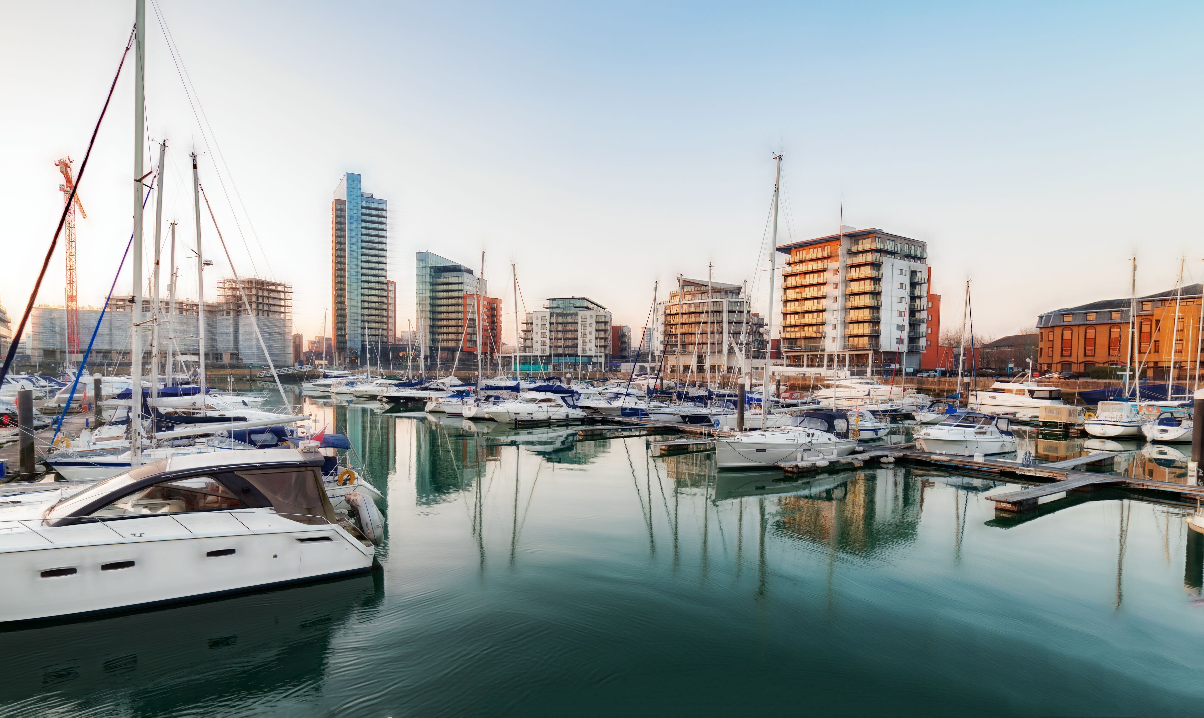 A marina with multiple boats docked, surrounded by modern buildings and cranes.