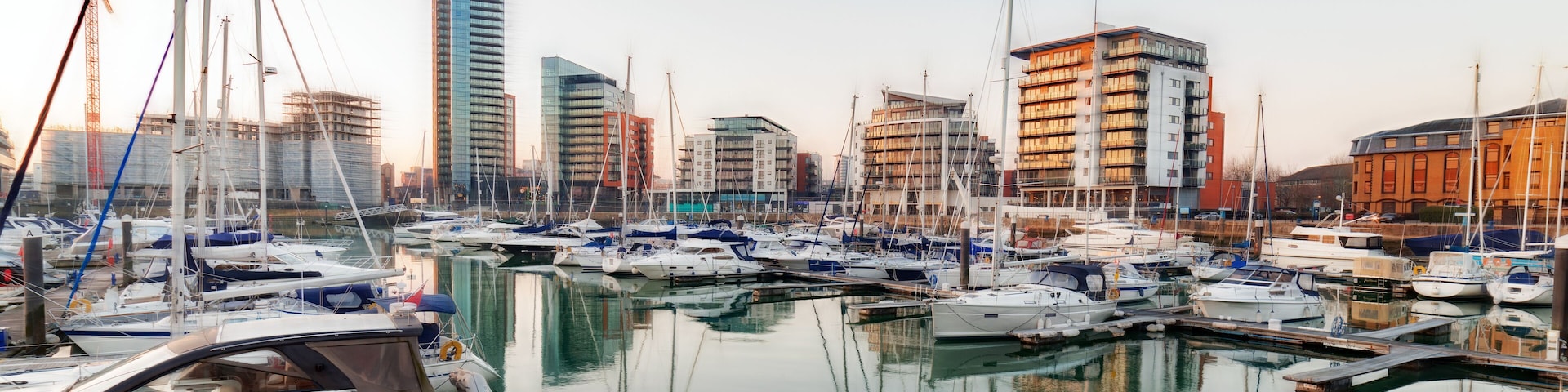 A marina with multiple boats docked, surrounded by modern buildings and cranes.