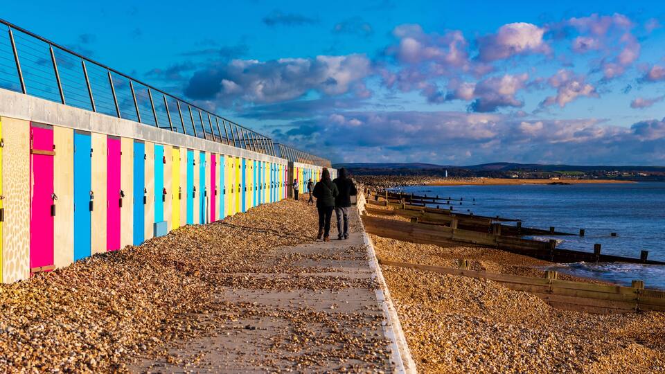 A pebbled beach with colorful wooden huts and a metal fence overlooking the sea.