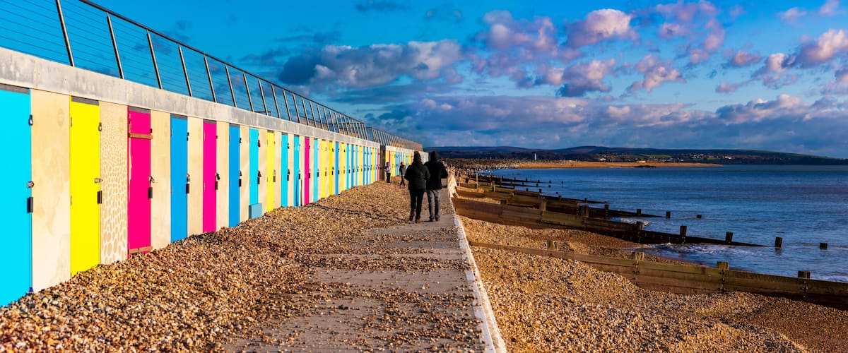 A pebbled beach with colorful wooden huts and a metal fence overlooking the sea.
