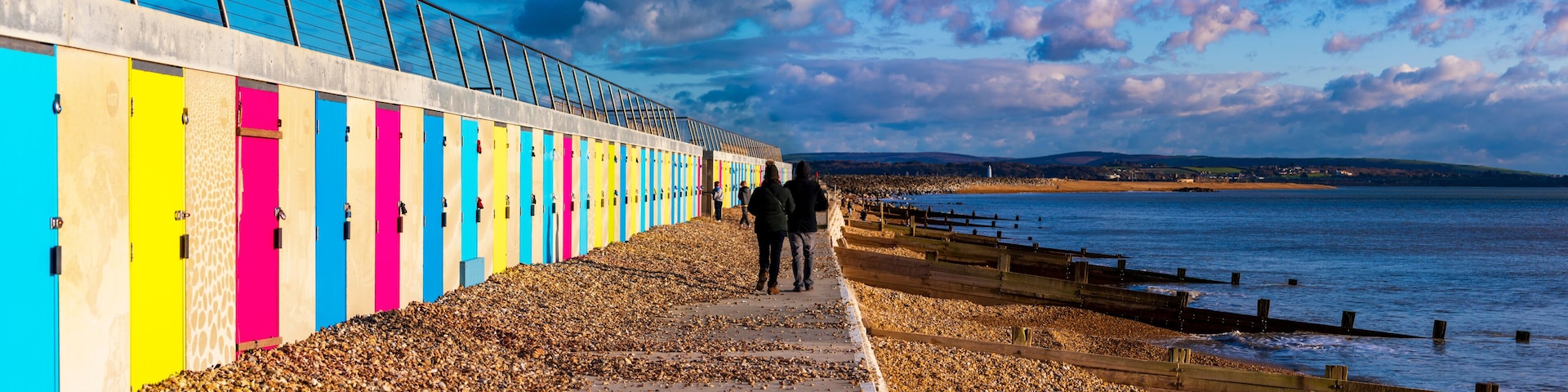 A pebbled beach with colorful wooden huts and a metal fence overlooking the sea.
