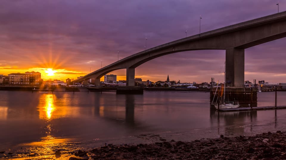A sunset over a river with a bridge and buildings in the background.