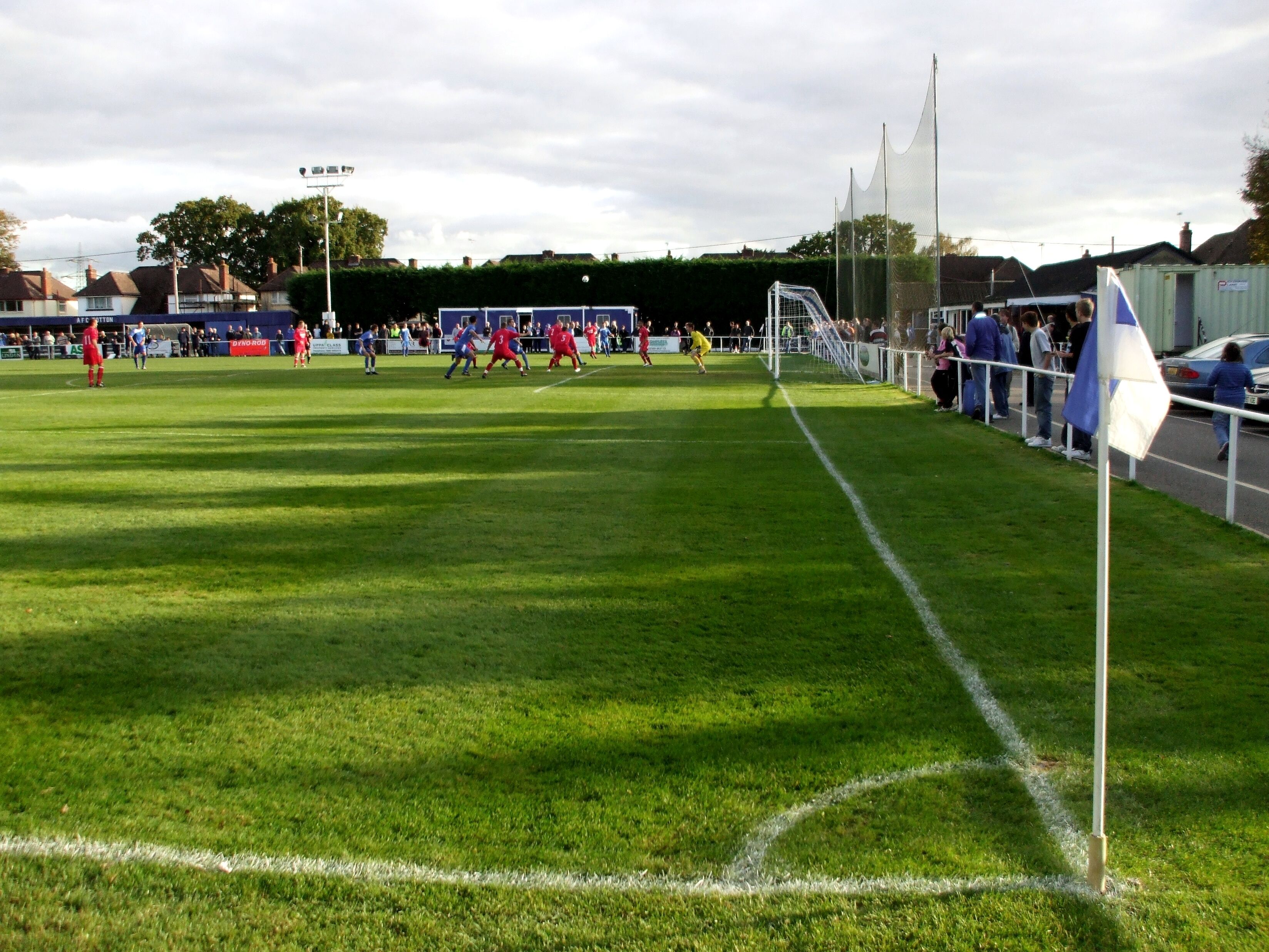 A soccer field with players in red and blue jerseys, a goalpost, and spectators in the background.