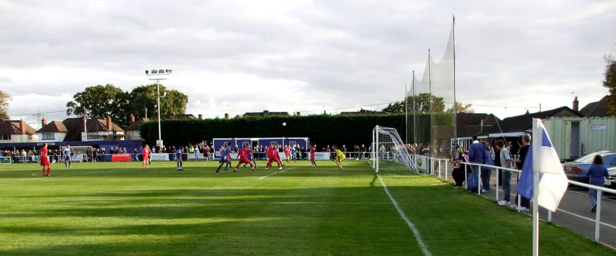 A soccer field with players in red and blue jerseys, a goalpost, and spectators in the background.