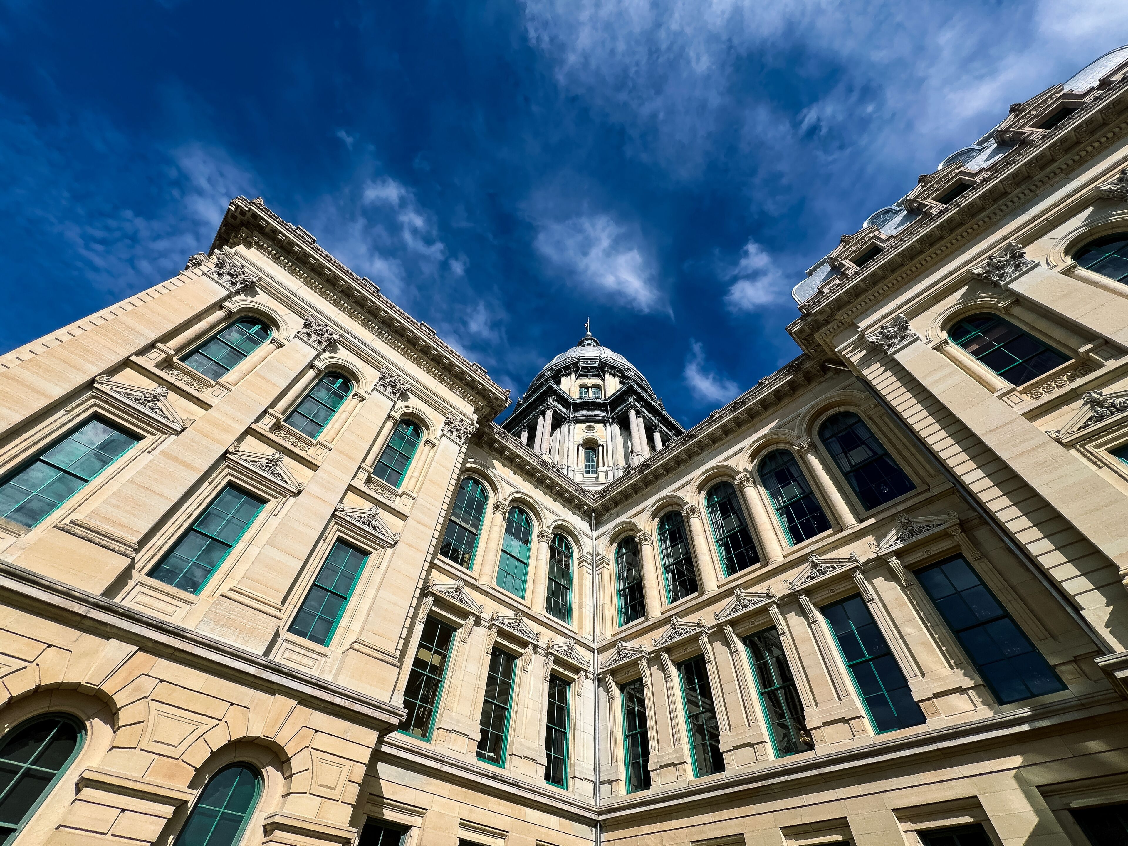 Rear views of the Illinois State Capitol Building in Springfield, Illinois, USA. Cloudy blue skies overhead. The US flag, State of Illinois Flag, and Black History Month Flag fly atop the dome.
