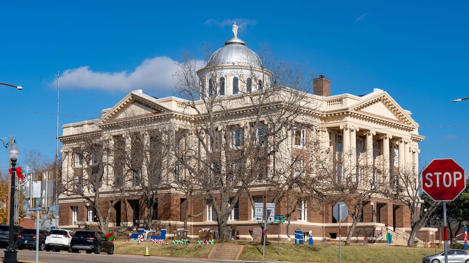 A large, historic building with a dome and columns, a stop sign, and a street with cars and a tree in front.