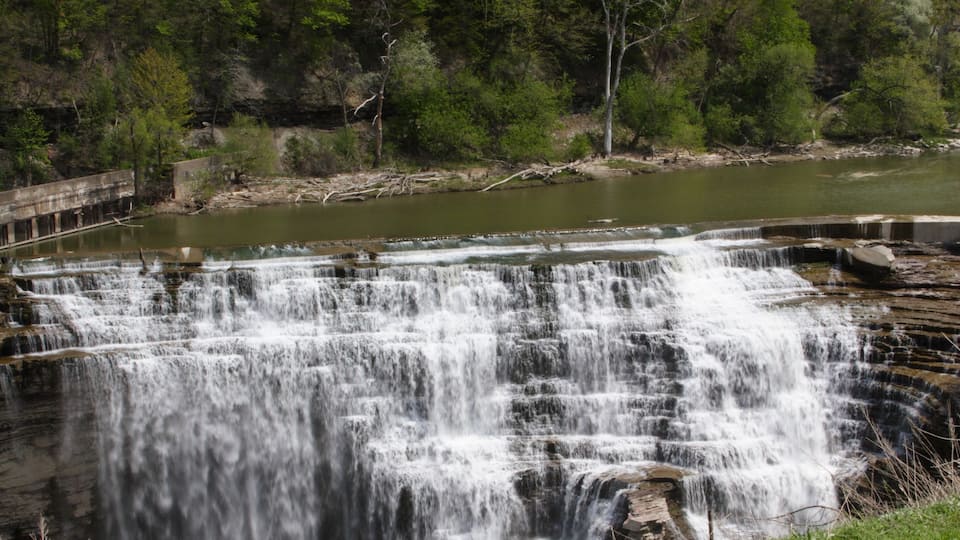 A multi-tiered waterfall cascading over rocks into a river, surrounded by greenery and trees.