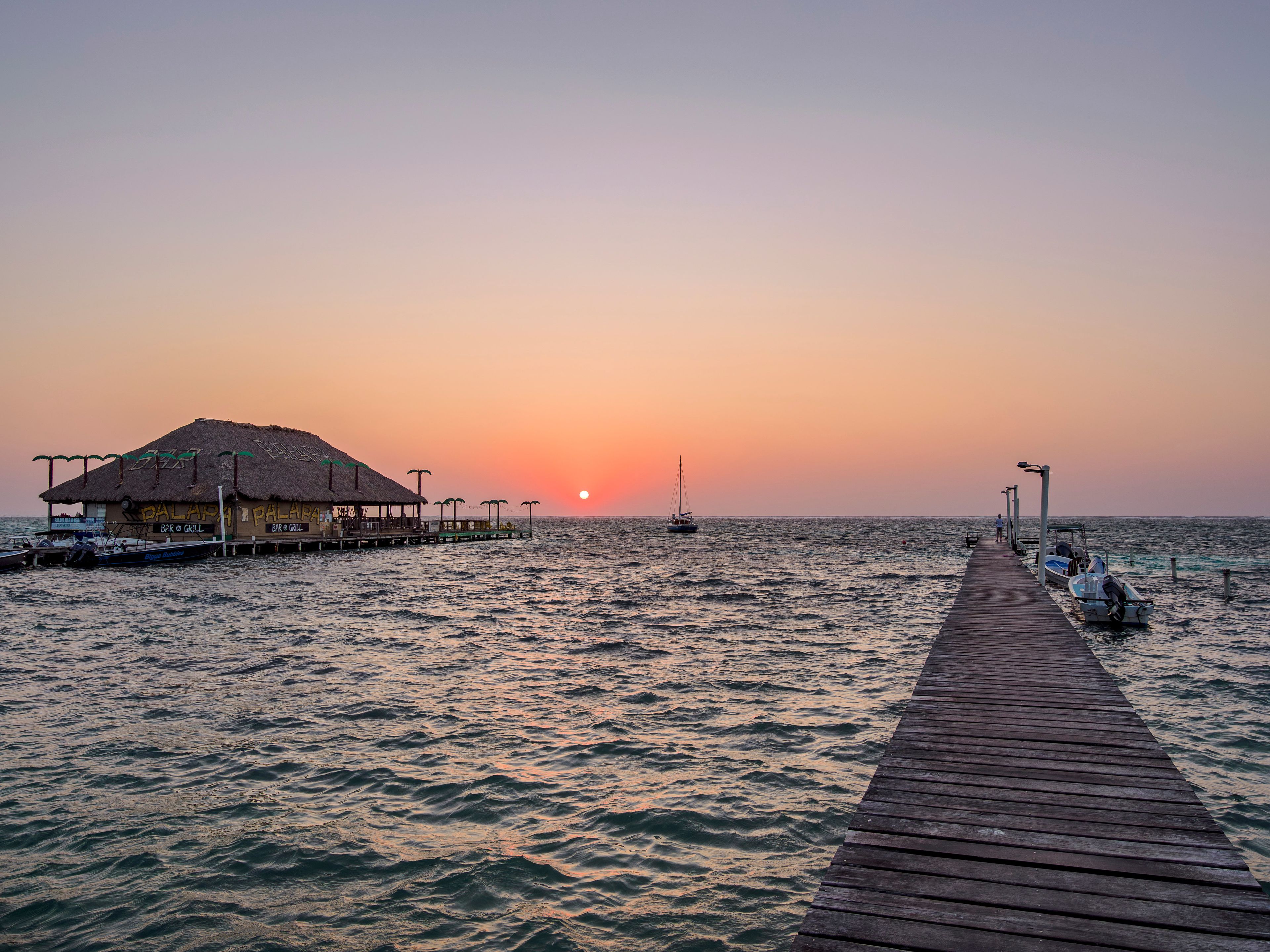 A sunset over the ocean with a pier leading to a thatched-roof building.