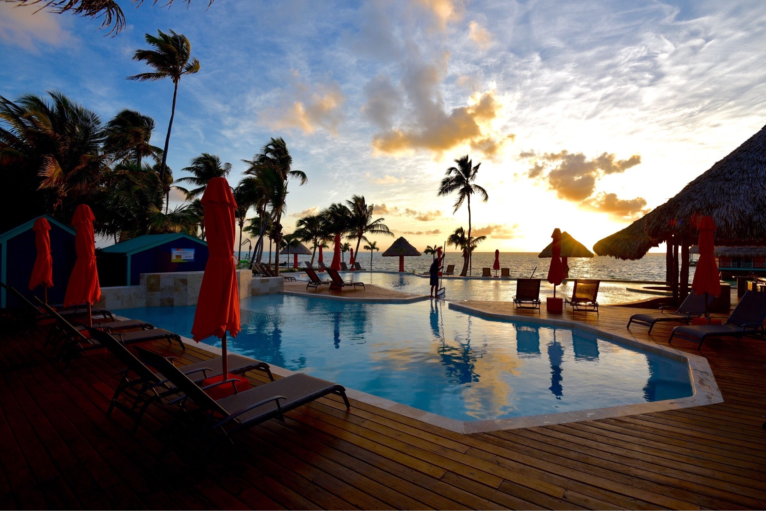 A poolside area with lounge chairs and umbrellas, palm trees, and a sunset over the ocean.