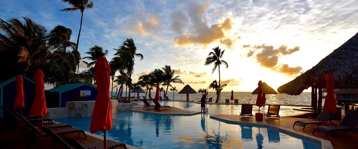A poolside area with lounge chairs and umbrellas, palm trees, and a sunset over the ocean.