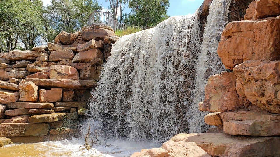 A waterfall cascading over a rocky ledge with a stone wall on the left and trees in the background.