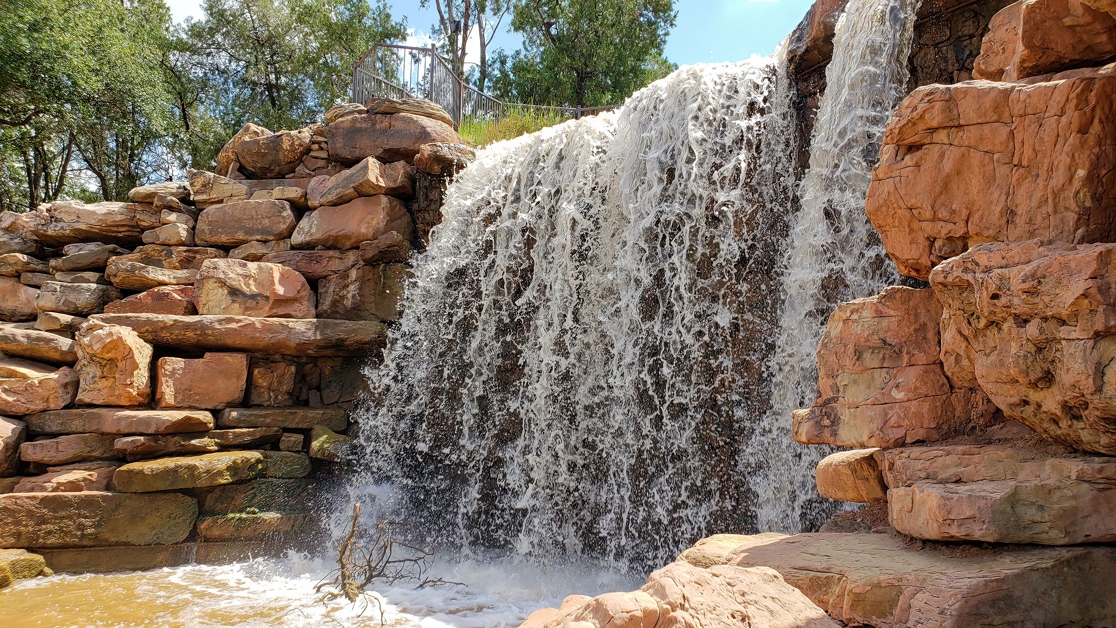 A waterfall cascading over a rocky ledge with a stone wall on the left and trees in the background.