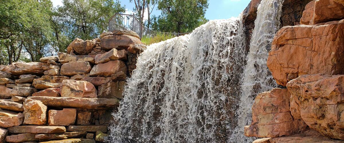 A waterfall cascading over a rocky ledge with a stone wall on the left and trees in the background.